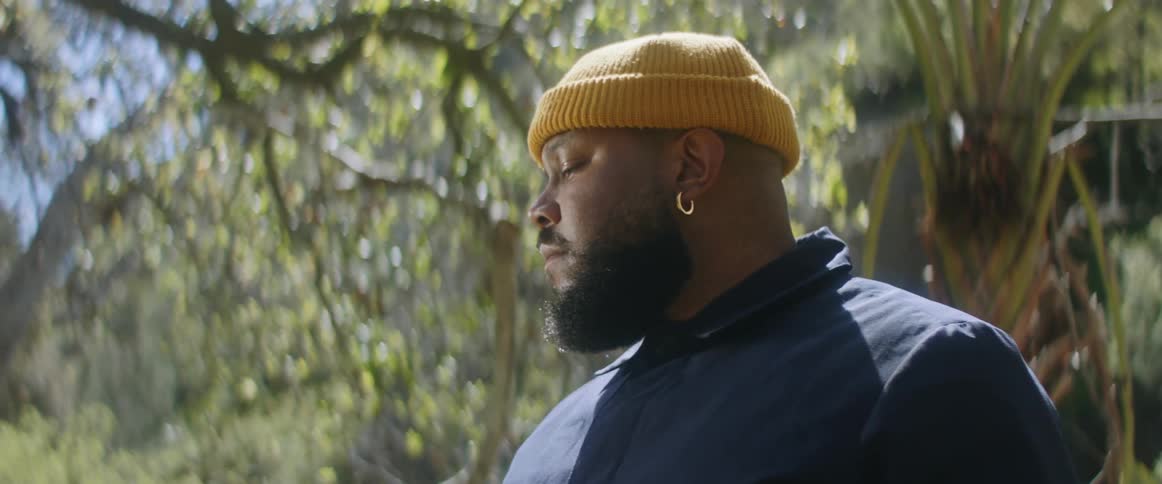 A black young man in the bush deep in thought he is wearing a yellow head warmer and has an earring in his ear