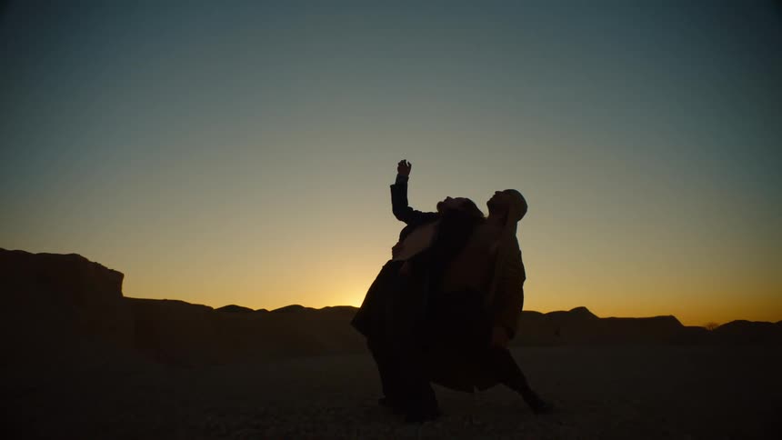 Two male dancers dance in arid plain with red sky and silhouetted mountains on the horizon