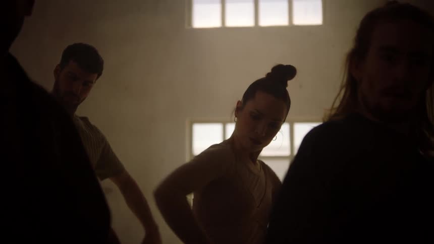 A woman dances vigorously in a darkened warehouse lit only by the light coming in through the windows along with a group of other dancers