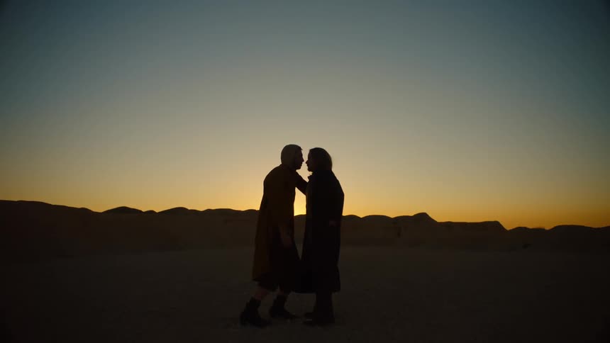Two men face each other while dancing in arid area with red sky and mountains on the horizon