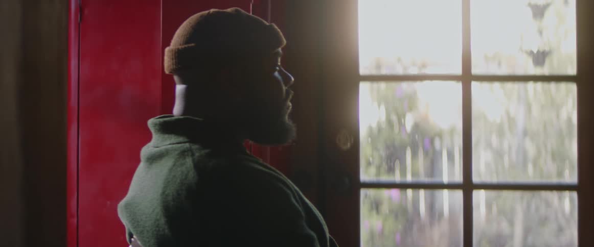 A man wearing a cap is sitting in a chair and he leans back as suns shines in the window