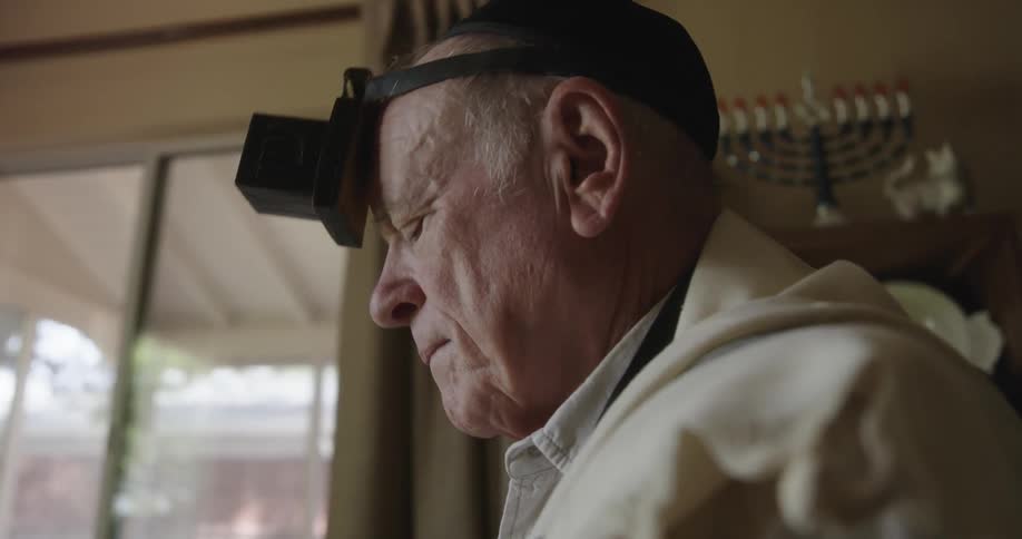 A jewish man wears a tefillin on his head and there is a menorah on the shelf behind him