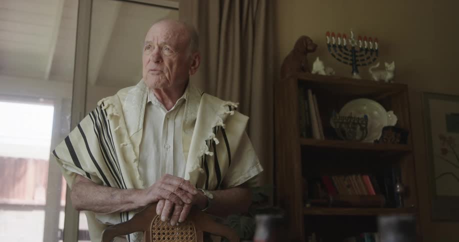 An older man is standing indoors in a dining room while praying during the day