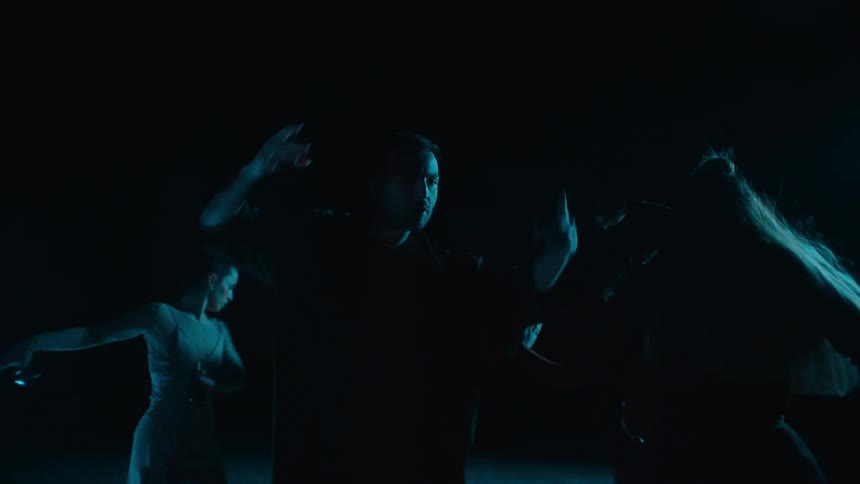 A group of performers dancing around and following the lead male performer while on a dark stage
