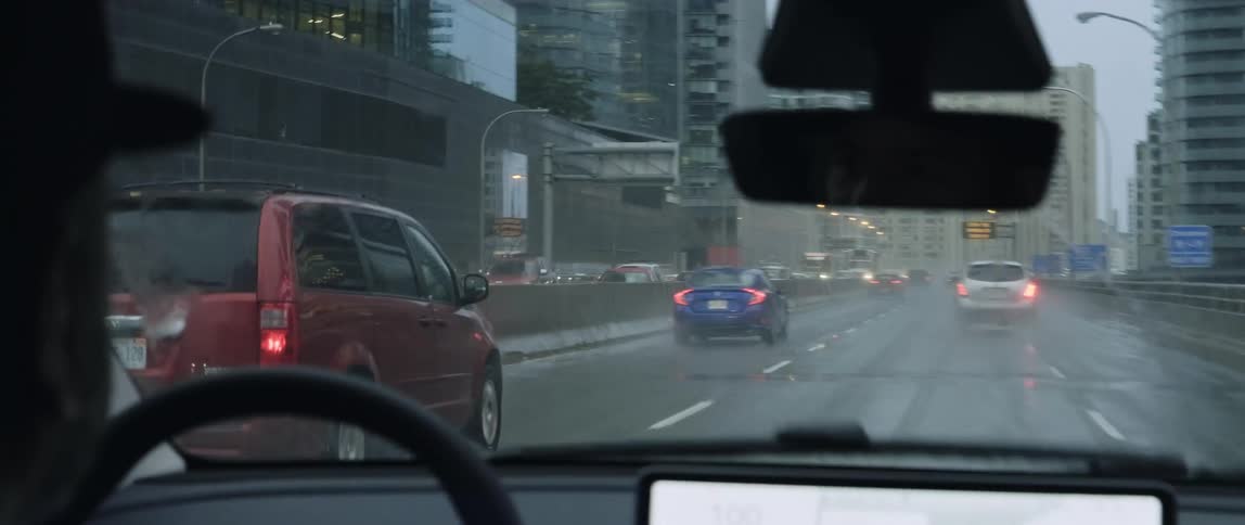 The inside of a vehicle being driven down the road by a person while it rains and the wipers clear the window