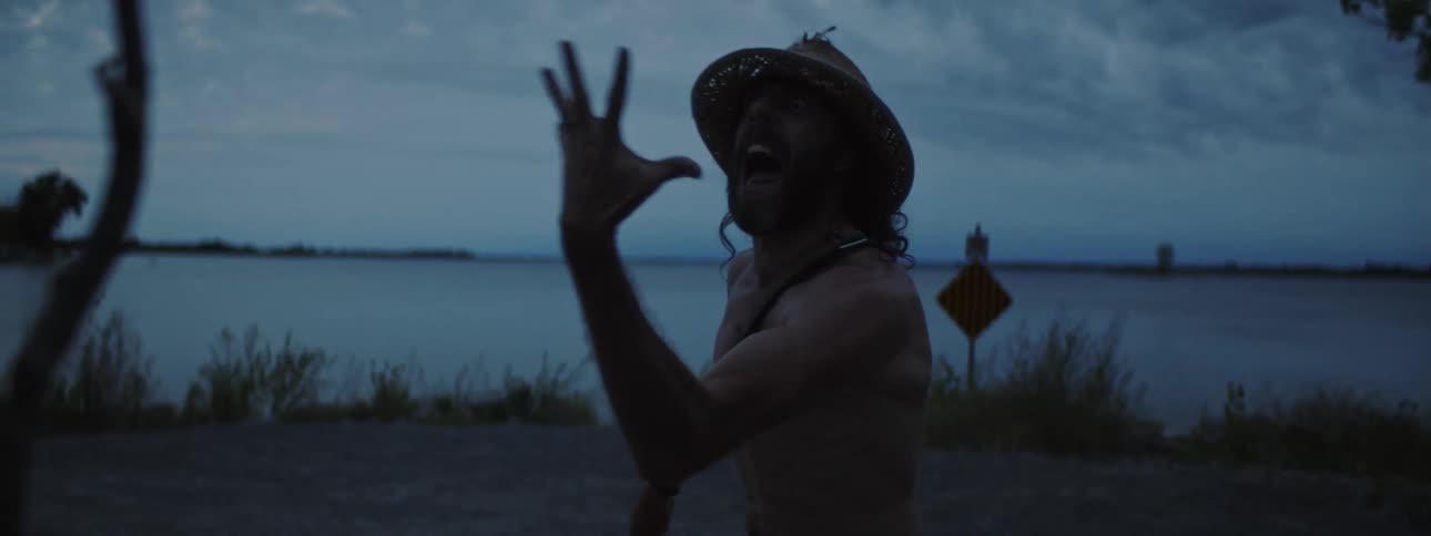 A man standing near a lake shore making aggressive and obnoxious movements and expressions at a person in front of him with a stick