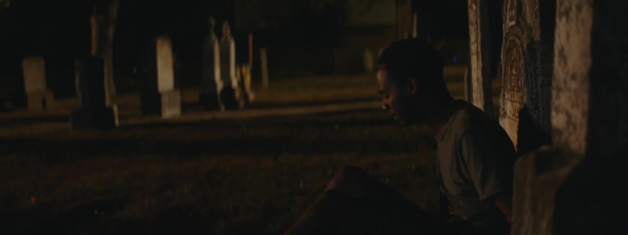 A young man looking exhausted sitting down in a cemetery against a head stone