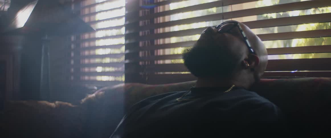 A man looking depressed reclining in a sofa in a darkened room lit only by daylight creeping through partially closed blinds