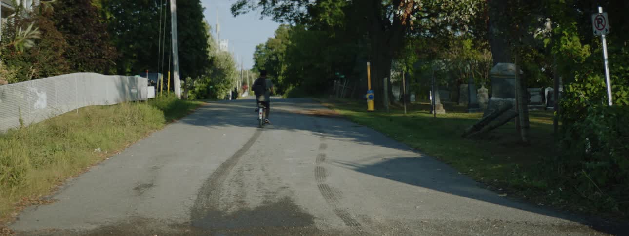 A teenage boy wearing a backpack is sitting on a bicycle and using his feet to push himself down a neighborhood street