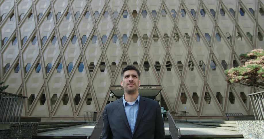 A man standing out front of a tall office building while smoothing over his hair with his hand