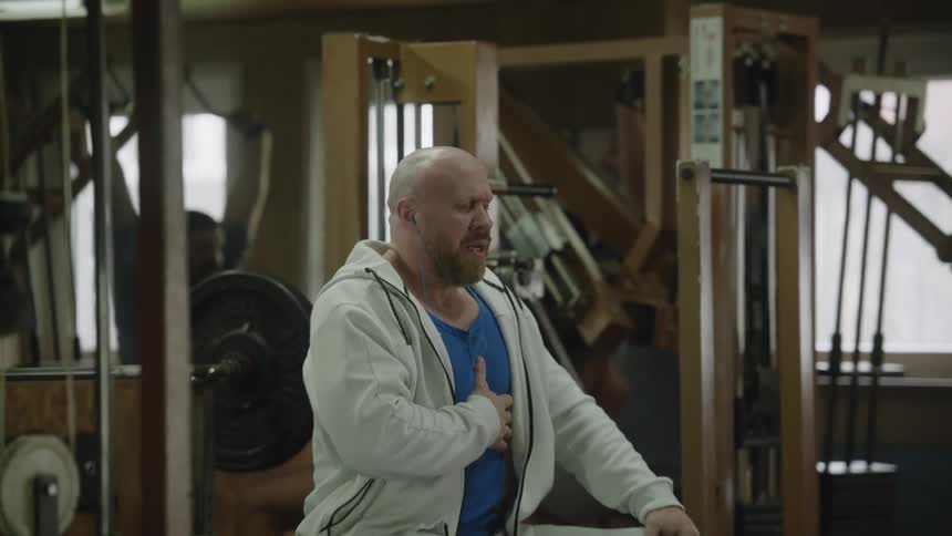 While others continue lifting weights in a weight room a man is sitting on a bench holding his chest and breathing heavily