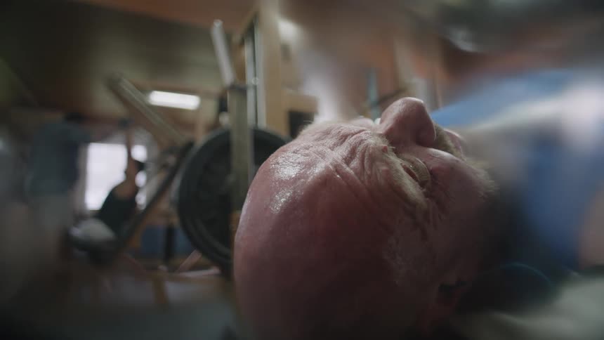 A bald man is in distress and gasps as he tries to bench press at the gym
