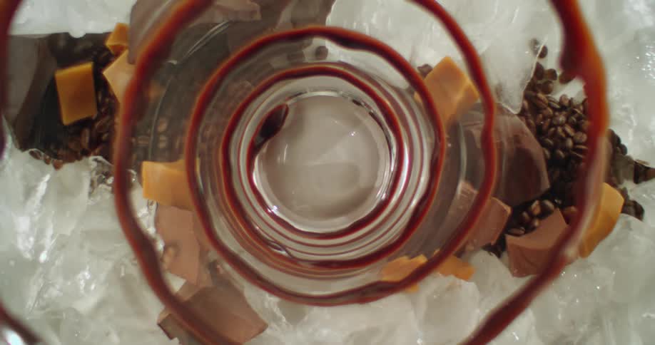 A close up of chocolate swirls in a glass cup as the cup gets bigger displaying caramel and fudge pieces around the glass