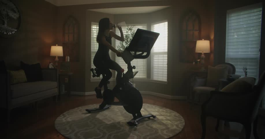 Woman with black top and leggings rides stationary bike while drinking water from bottle in the living room