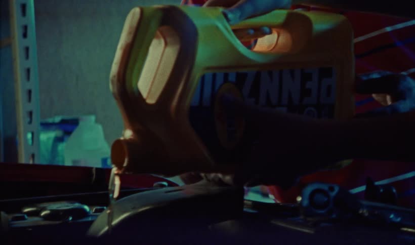 Two people pouring a yellow oil canister over a vehicle motor in a garage