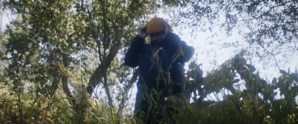 A man putting on a yellow beanie cap while standing outside near trees