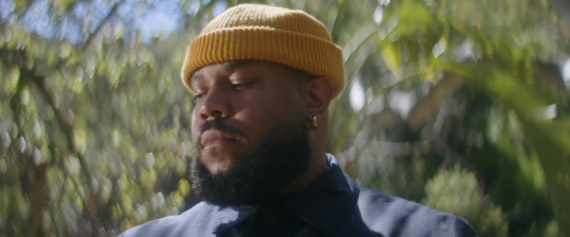 Man with beard earrings and yellow beanie stares down at camera in the forest