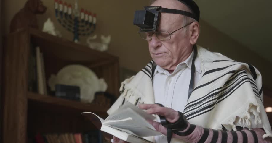 An old man standing in his house looking through a book while wearing a black strap on his head and arm