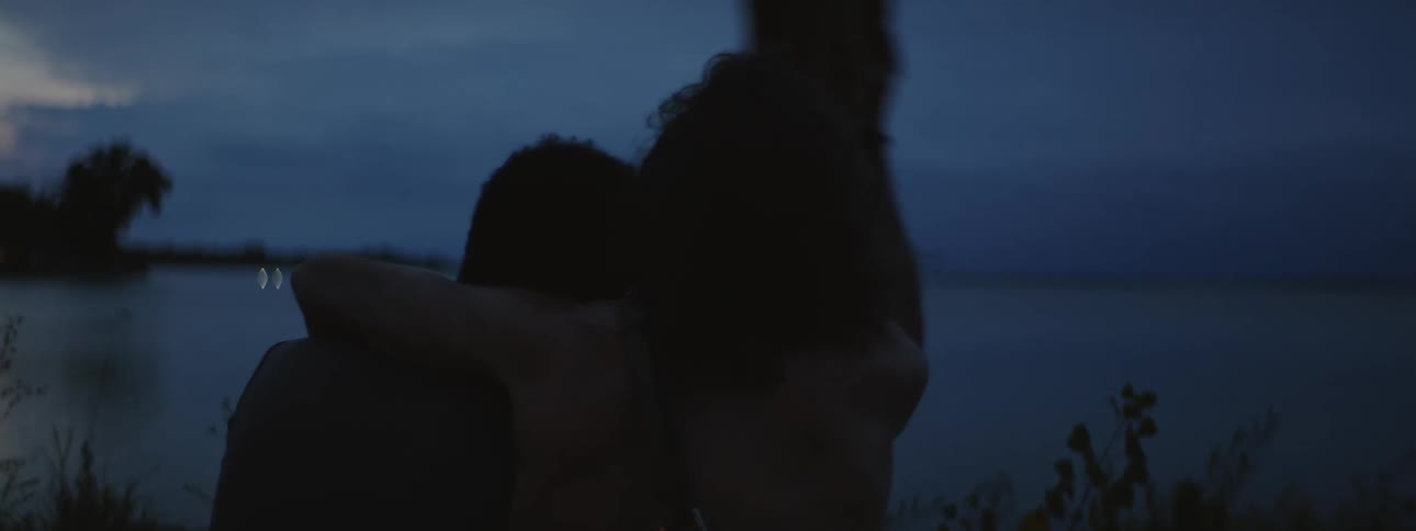 Two men sitting together at sundown at the lake one puts his arm around the other and puts his hat on his head