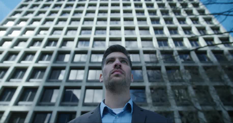 Man with blazer jacket and blue shirt stands in front of high rise office building