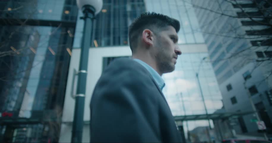 A man walking down the city sidewalk near light poles and trees with large buildings in the background