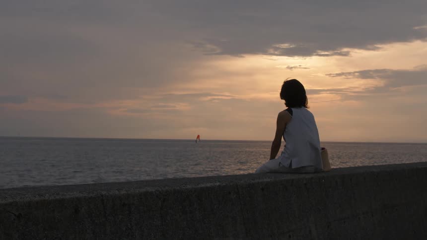 A person sitting on a concrete wall looking out at the ocean as a boat sails on the sea in the distance