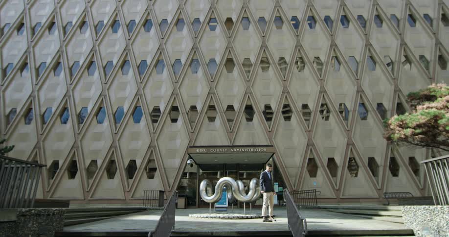 A man walking around in circles outside of a large building with a unique sculpture in front