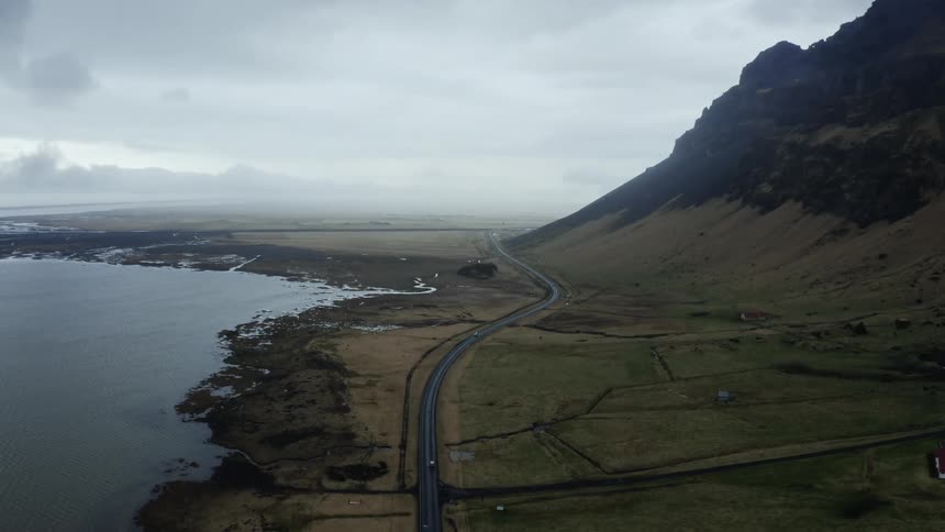 An ocean with a long tile road running alongside it cars are moving along the road situated close to a picturesque hill