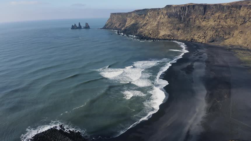 Waves crash into the coastline in front of a cliff face broken by a pier
