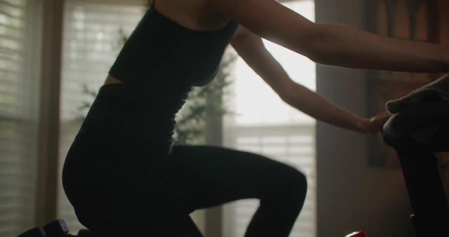 A woman is riding an exercise bike and she moves in position to pedal hard