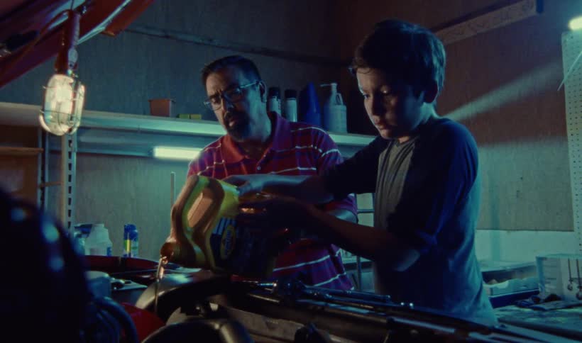 A man instructs a boy as the boy pours oil into the engine of a vehicle inside a garage