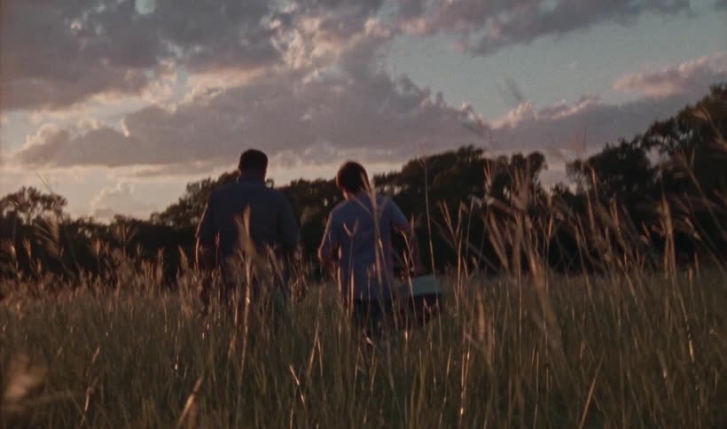 A man and a boy walk together through a field while carrying fishing equipment