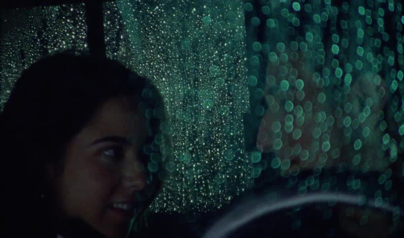 Woman and man gaze at each other while sitting at night in vehicle with wet windows
