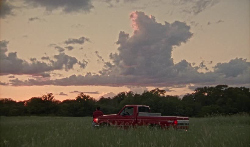 A man standing against his red truck outdoors in a field lined with trees at dusk