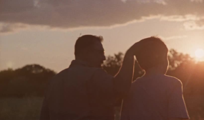 A man and his young son are carrying fishing poles as they walk across a field and the man rubs his sons head as the sun shines brightly over the trees