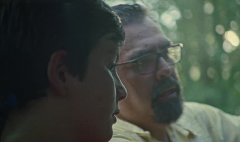 A father is teaching his teenage son how to drive while his son holds the wheel