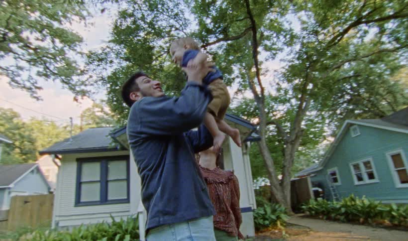 Parents hold their son while standing in yard as cyclist rides along road