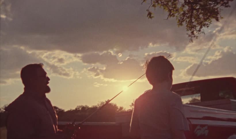 A father and son are retrieving fishing poles and a tackle box from the back of a red pickup truck
