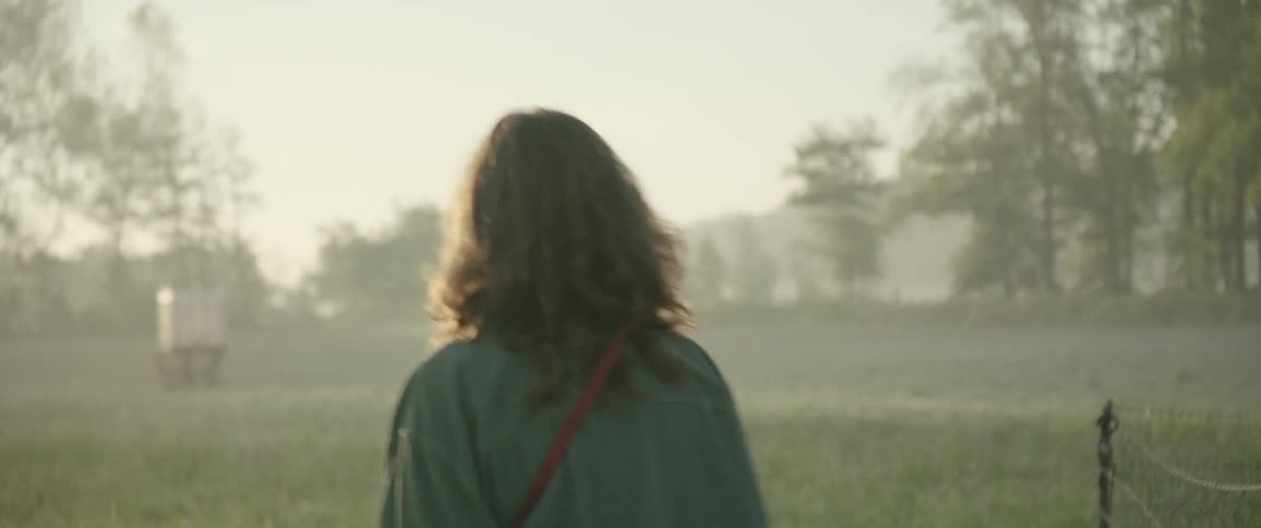 The sun shines brightly upon a teenage girl walking in a foggy field with mountains in the distance