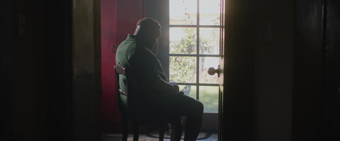 Man wearing a cap sits on a chair inside a dimly lit room near a window at daytime