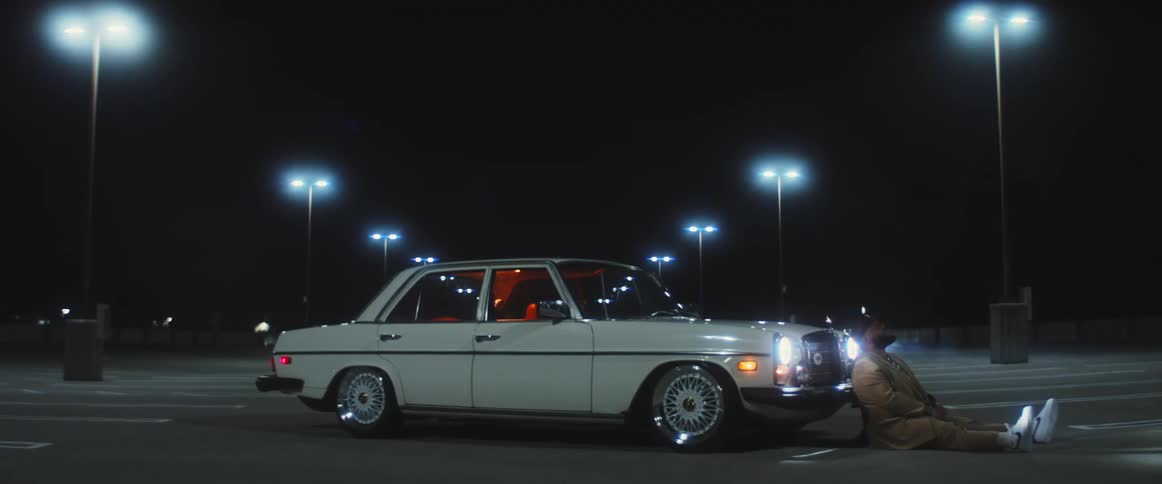 Man sitting on the ground reclined on the bumper of a car parked on a parking lot at night