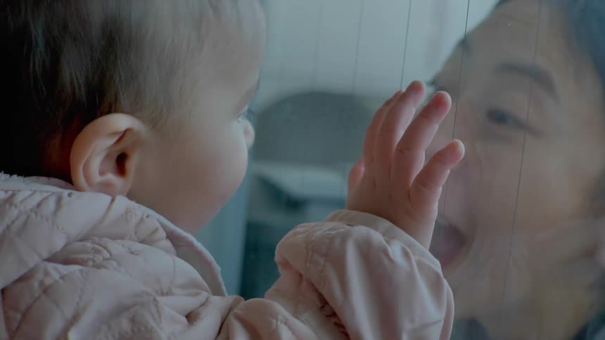 A woman makes faces at a young toddler through a glass window