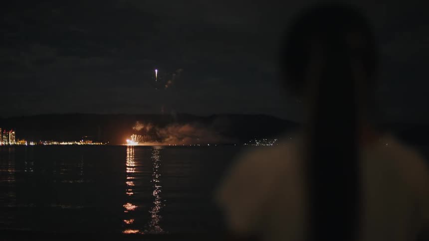 Back of a woman looking at fireworks show over a lake at night