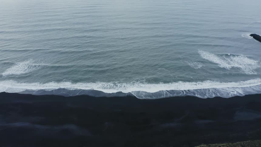 Aerial of gentle waves crashing on shoreline at a black sand beach