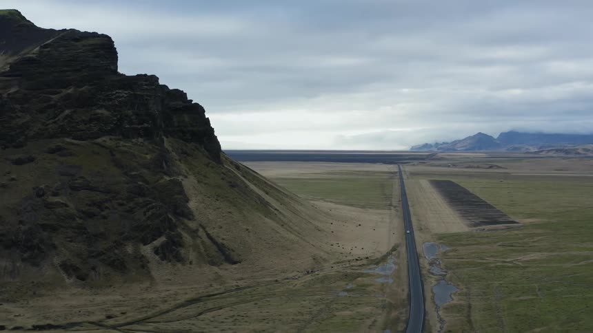An aerial view of a road running through a plains landscape near a rocky peak with mountains in the background