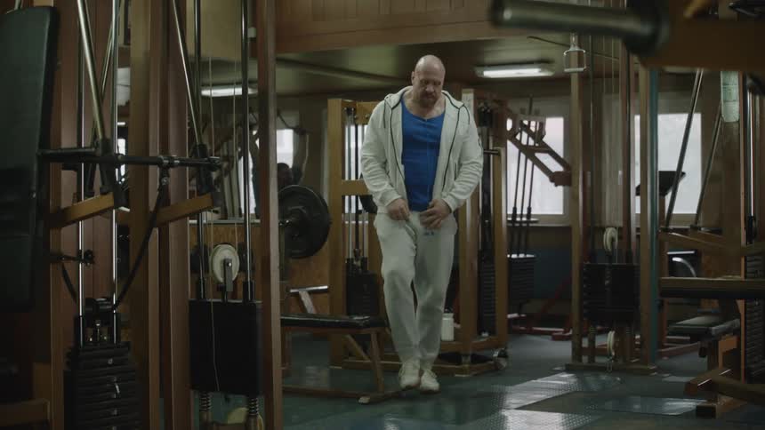 A man in a gym wearing all sweats and a blue undershirt sitting down on a weight bench getting ready to lift