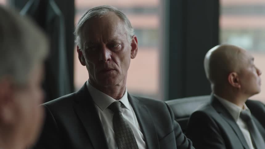 An older man looks weak and worried while sitting in a meeting and adjusting a hearing aid