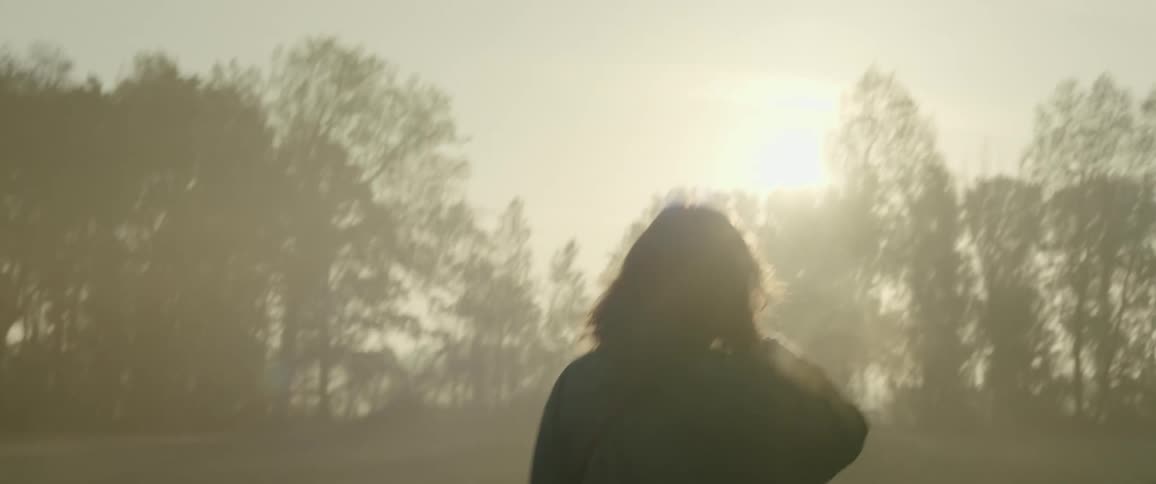 Woman silhouetted by sun walks across misty field
