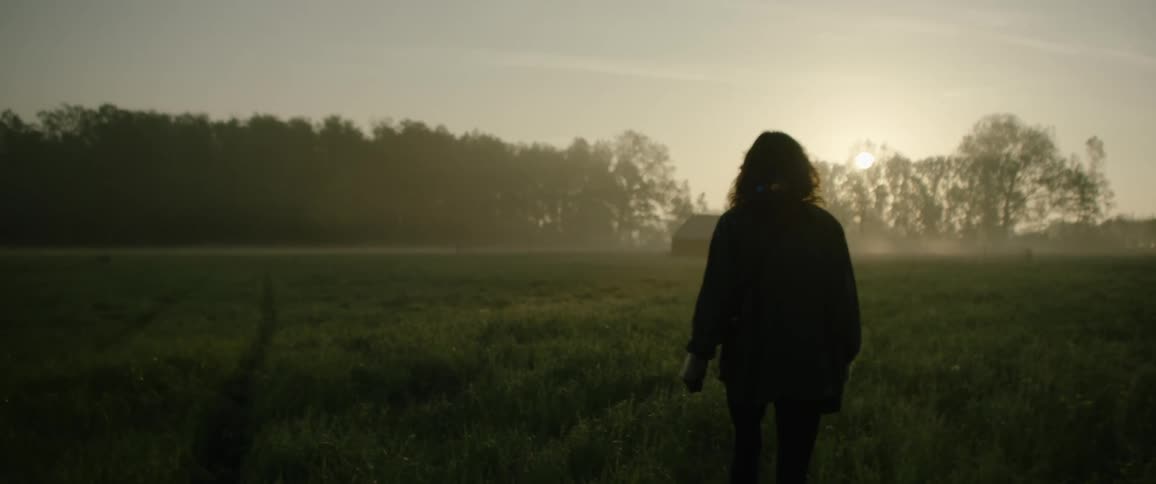 A person walks across a grassy field towards a barn at dusk with trees in the background