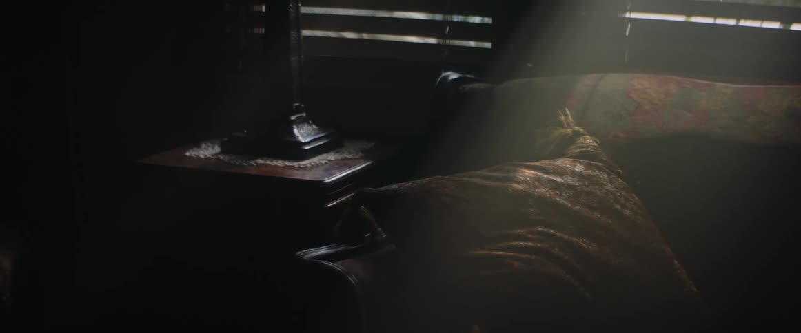 A close up of a couch and pillow next to an end table with a lamp stand on it with light peaking through the shades